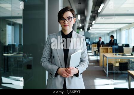 Portrait of beautiful young woman with short hair in formal suit holding digital tablet and looking at camera standing at office Stock Photo
