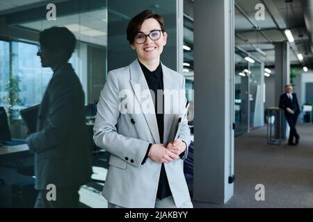 Portrait of young successful female leader in formal suit smiling at camera standing at modern office Stock Photo