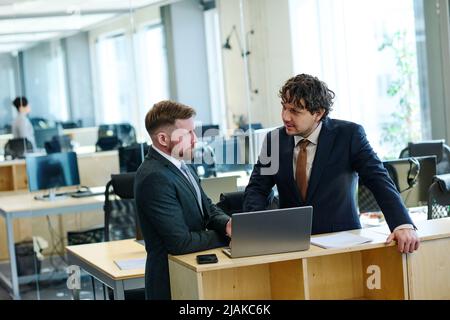 Two colleagues in formal wear standing at table at office and talking while using laptop Stock Photo