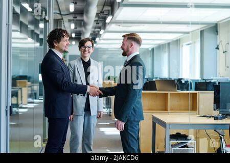 Business partners in formal suits greeting each other before meeting with young businesswoman standing near by them and smiling Stock Photo