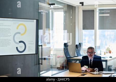 Mature manager in suit sitting at workplace with laptop and using his mobile phone, he working at modern office behind glass wall Stock Photo