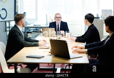 Senior manager in suit talking to partners at table with laptops during meeting at meeting room Stock Photo