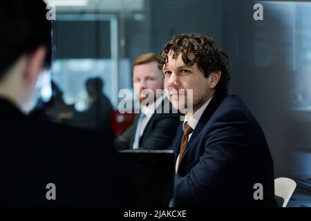 Young businessman in dark suit listening to speaker while sitting at table with his colleagues at meeting Stock Photo