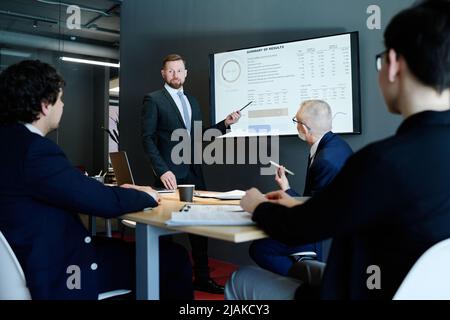 Young businessman in suit pointing at display screen with charts and performing with his presentation for colleagues at meeting room Stock Photo