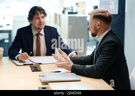 Two colleagues in formal suits discussing business plan in team at table during meeting at office Stock Photo