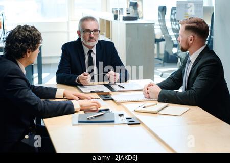 Group of serious businessmen in suits sitting together at table at boardroom and planning work in team Stock Photo