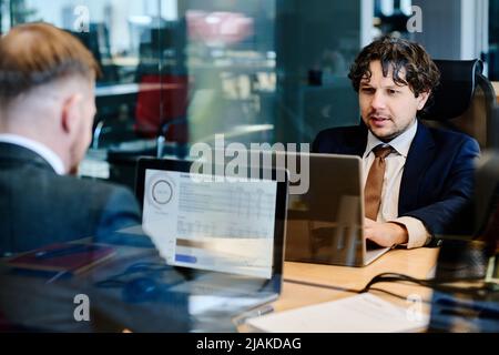 Two businessmen sitting at table with laptops opposite each other behind glass wall, they examining financial marketing Stock Photo