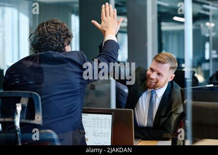 Two business colleagues in formal suits giving high-five to each other and celebrating success at their workplaces with laptops Stock Photo