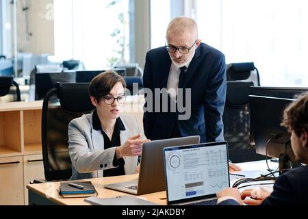 Young businesswoman sitting at her workplace and pointing at laptop, she consulting with senior manager Stock Photo