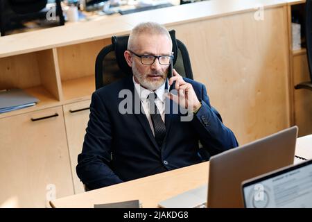 Serious senior businessman in dark suit sitting on chair at workplace in front of his laptop and talking on mobile phone Stock Photo