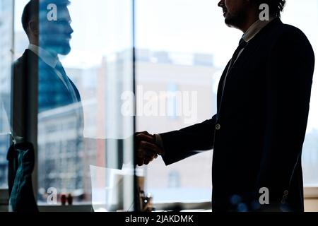 Two businessmen in suits shaking hands to conclude a deal at office standing behind glass wall Stock Photo