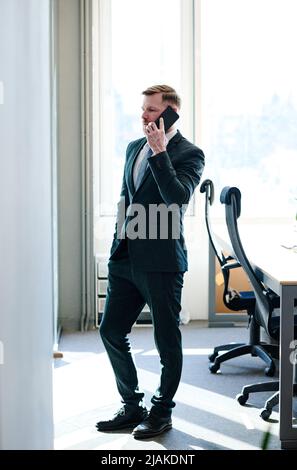 Young businessman in formal suit talking on mobile phone while standing at office Stock Photo