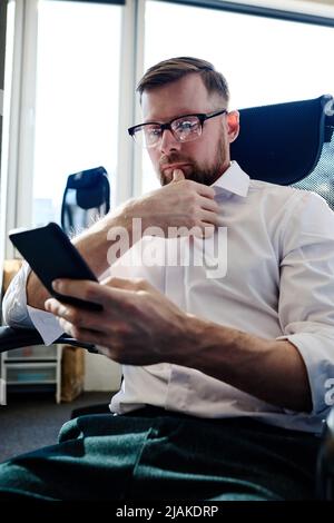 Young bearded businessman in eyeglasses sitting at office chair and working online on mobile phone Stock Photo