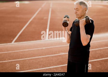Image of athletic old man in tracksuit doing exercise while working out in morning. Man with dumbbells outdoors. I feel strong and healthy. Start your Stock Photo