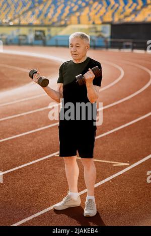 Senior man doing exercise with dumbbells in black sportswear outside. Mature concentrated man doing sports to keep fit. Stock Photo