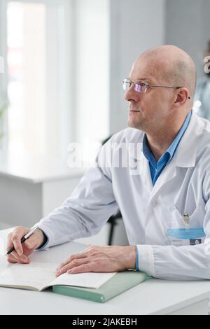 Mature doctor in eyeglasses and white coat sitting at desk with notebook during training, he focused on studying Stock Photo