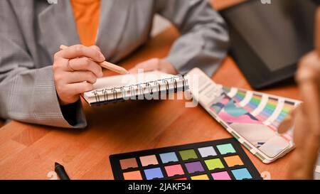 Professional female graphic designer taking lists on her spiral notepad at her office desk. colour palette and colour checker board on table. close-up Stock Photo