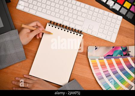 Graphic designer office desk with a female hand writing something on blank spiral notepad. top view image Stock Photo