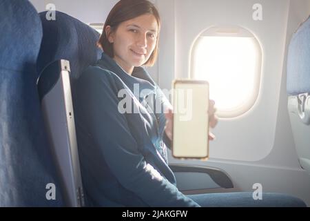 Young beautiful woman sitting at window of plane during the flight. shows smartphone monitor screen, mock-up Stock Photo