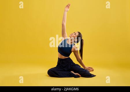 Front view of flexible, pretty woman sitting in pose of yoga in studio. Slim girl in black activewear posing with arm, stretched in air, isolated on yellow studio background. Concept of yoga. Stock Photo