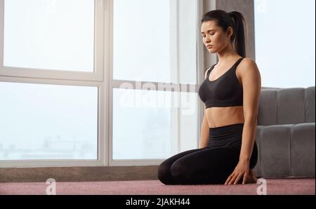 Side view of serious peaceful girl sitting in yoga pose with closed eyes and meditating next to window. Beautiful brunette woman practicing yoga on floor, in bright studio. Concept of meditation. Stock Photo