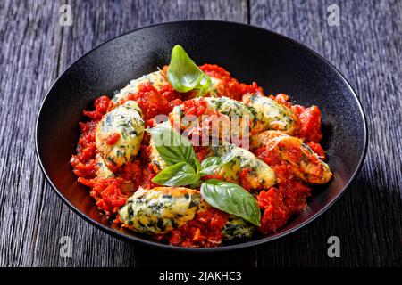 Malfatti, Italian spinach ricotta dumplings  in tomato sauce with herbs and grated parmesan cheese in black bowl on dark wooden table Stock Photo