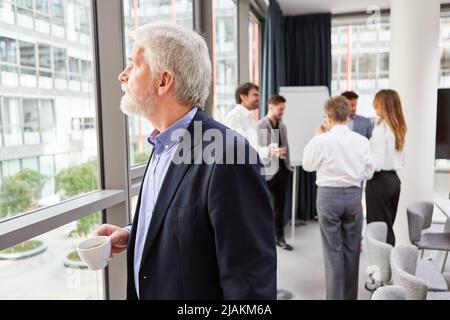Manager standing by the window in the office drinking a cup of coffee with team in the background Stock Photo