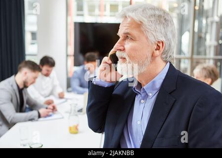 Experienced manager talking on mobile phone in meeting with team in background Stock Photo