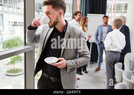 Young business man drinking a cup of coffee at the window in the office with team in the background Stock Photo