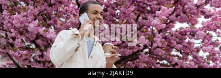 smiling african american man holding takeaway drink and talking on smartphone near blooming cherry tree, banner Stock Photo