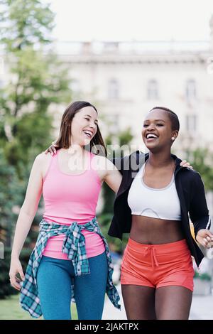 Cheerful smiling friends in sportswear walking after a sport session in the city discussing and having fun. Multiethnic women having a fitness workout jogging. Stock Photo