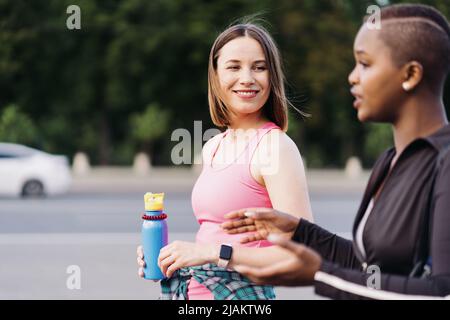 Cheerful smiling friends in sportswear walking after a sport session in the city discussing. Multiethnic women having a fitness workout jogging. Stock Photo