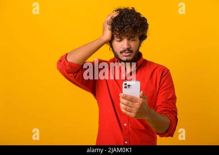 Thoughtful bearded man scratching head holding smartphone in hand, contemplating about software updating, choosing suitable tariffs. Indoor studio shot isolated on orange background Stock Photo