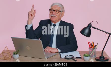 Eureka. Inspired senior business man raises finger came up with creative plan feels excited with good idea inspiration motivation using laptop. Elderly grandfather freelancer at office pink background Stock Photo