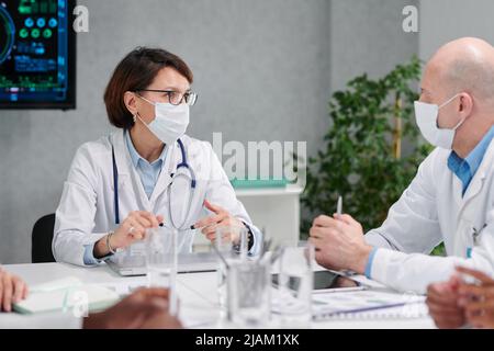 Two colleagues in masks and white coats discussing together at table during meeting at hospital Stock Photo
