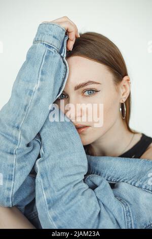 Vertical of calm, serious dark-haired woman looking at camera, wearing denim jeans jacket. Hugging herself. Close-up Stock Photo