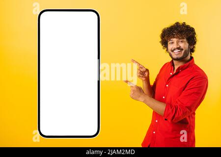 Playful handsome curly indian bearded man points fingers at huge smartphone with empty blank screen isolated on yellow, happy latin guy presenting mobile app, showing best deal, copy space, template Stock Photo