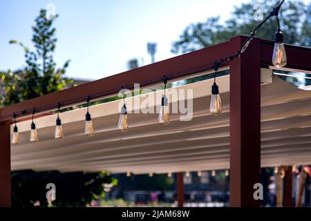 textile awning in the backyard wooden frame gazebo with a garland of strings of retro edison light bulbs of lamps lighting glowing with warm light on Stock Photo