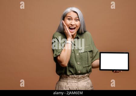 Portrait of the shocked surprised woman holding tablet and looking at the camera with big astonished eyes. Indoor studio shot isolated on beige background Stock Photo