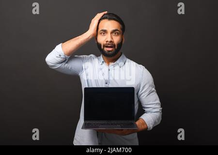 Discouraged Indian bearded freelancer guy holding and showing laptop computer with empty screen isolated on black background, worried dark-haired 30s multiracial man holding head in despair Stock Photo