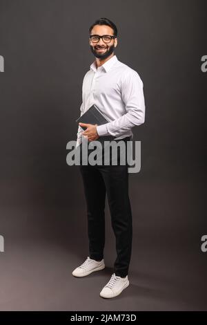 Full length of happy Indian bearded man in smart casual wear and stylish glasses standing isolated on black background and carrying digital tablet Stock Photo
