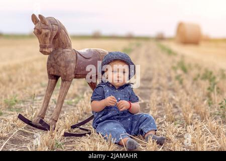 happy and smiling cute baby girl on field of wheat with rocking horse. Stock Photo