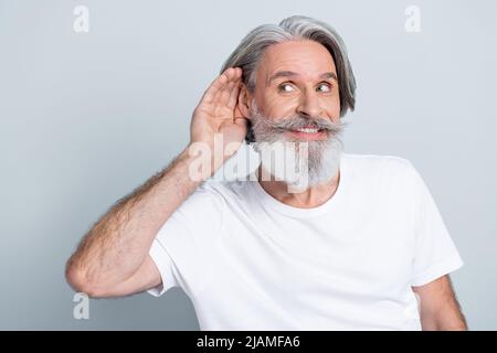 Photo of funny curious man pensioner dressed white t-shirt hearing secrets looking empty space smiling isolated grey color background. Stock Photo