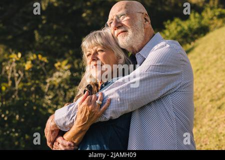 Romantic senior couple embracing each other while standing in a park. Elderly couple enjoying the sun in each other's arms. Mature couple spending qua Stock Photo