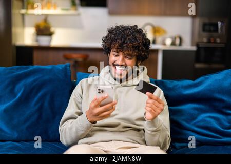 Smiling Indian man holding banking card and smartphone, making purchases or ordering online. Mixed-race guy using mobile app for booking or renting, paying online with credit card sitting on the coach Stock Photo