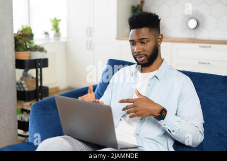 Smiling young bearded man sitting on the couch with laptop on his lap, using dating app, video-calling girlfriend online, gesturing and talking, blogger streaming his daily life Stock Photo