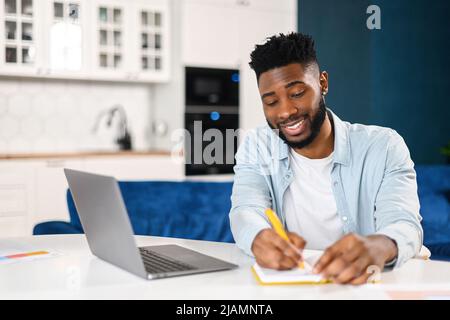 Modern bearded man working with laptop in home office. Multiracial male in smart casual shirt watching webinar and writing down, taking notes with a pen, planning schedule Stock Photo