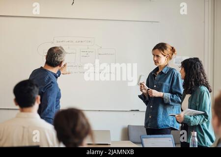 Male teacher explaining diagram to students while pointing at whiteboard in classroom Stock Photo