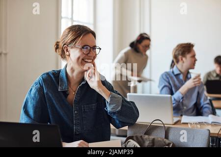 Happy female student with eyeglasses looking away while sitting in classroom Stock Photo
