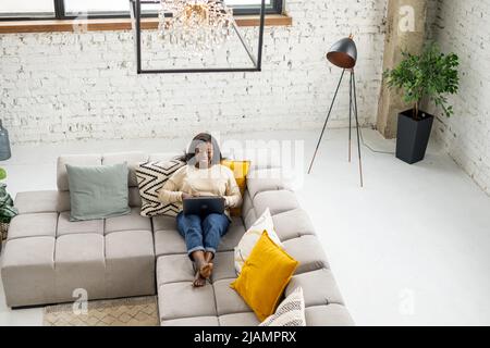 High angle view of the cheerful lady spend leisure time in network, web surfing. Relaxed multiracial woman sitting on the couch and chatting online on the laptop, answering email Stock Photo